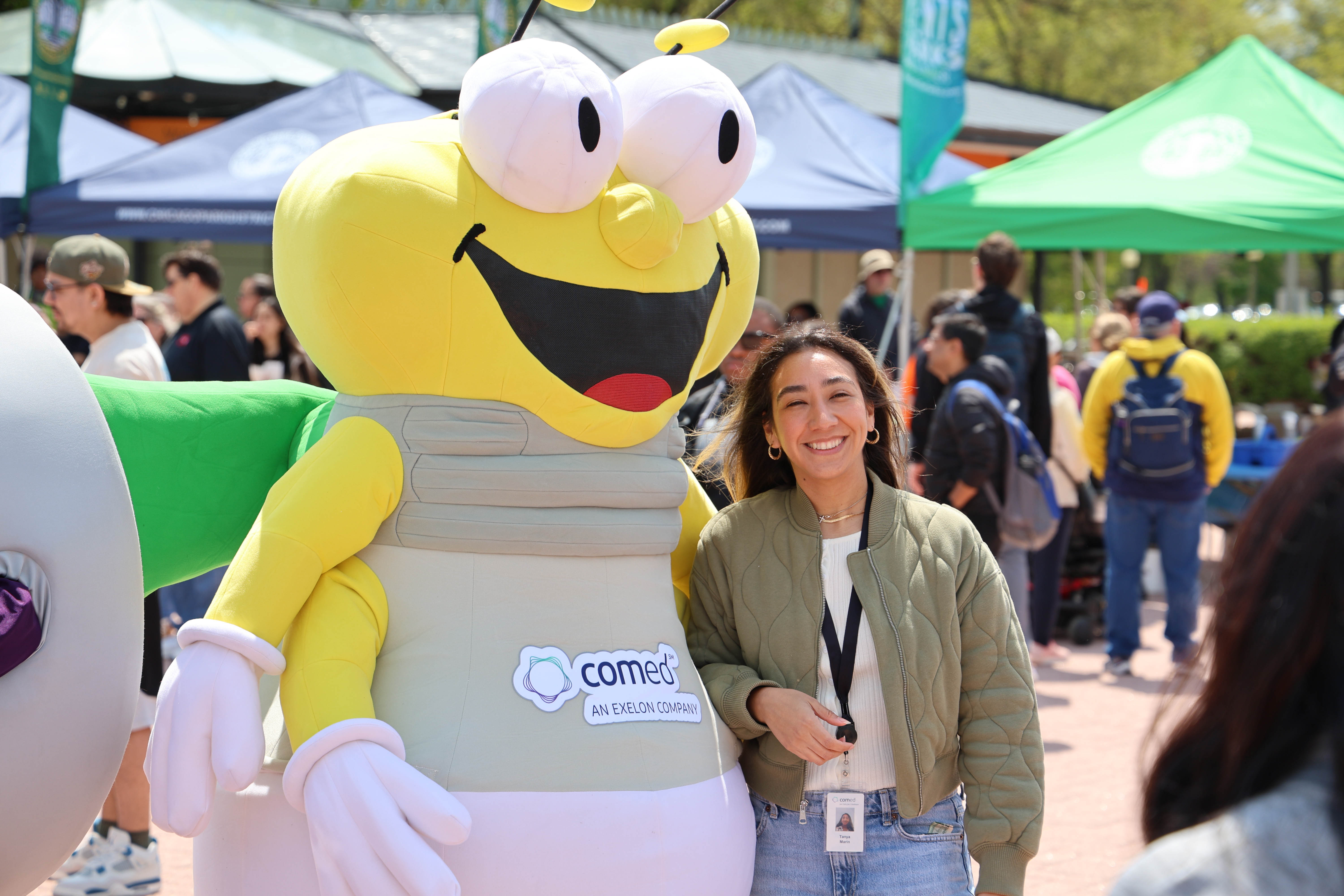 A woman smiles beside a large, friendly bee mascot at an outdoor event. The mascot wears a ComEd logo.
